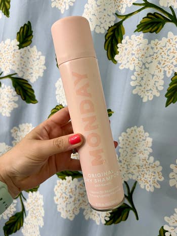 Person holding a tin  of Monday Original Dry Shampoo against a floral-patterned background