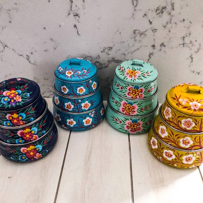 four hand-painted, three-tiered bento boxes in navy blue, light blue, aqua, and yellow, all with floral designs 