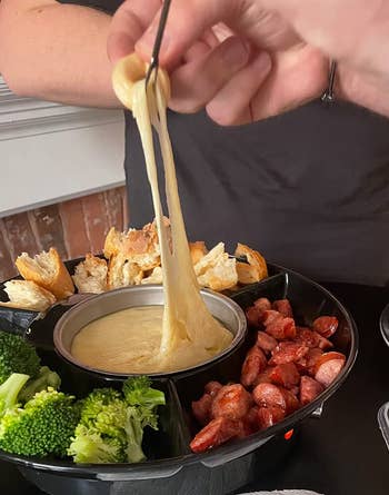 Person dipping breadstuff  into a food  fondue cookware  surrounded by bread, broccoli, and sausage