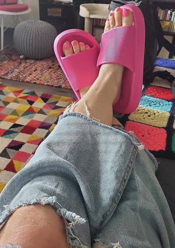 Person relaxing with feet on a table, wearing pink slides and ripped jeans, colorful rug and room interior visible