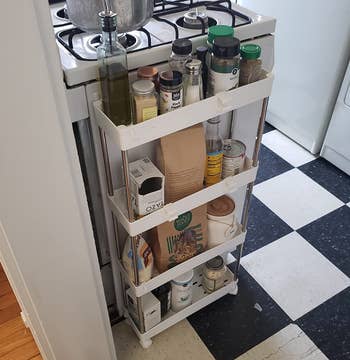 A narrow, rolling storage cart with four shelves, filled with various kitchen items like spices and oils