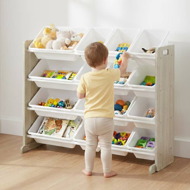 Toddler organizing toys in a white multi-bin storage unit, including books, plush toys, and colorful plastic items. Suitable for tidy playrooms