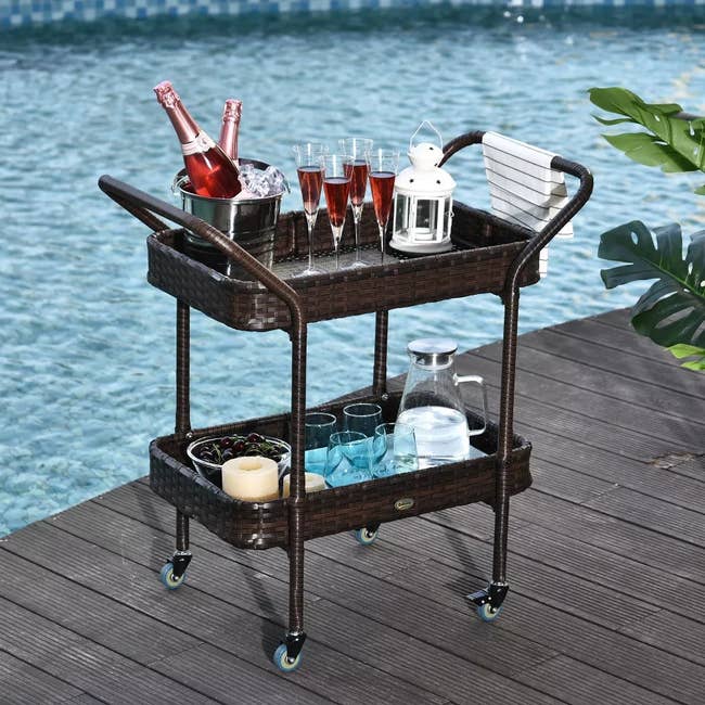 Wicker outdoor bar cart by a pool with wine bottles, glasses, a lantern, candles, a bowl of grapes, and a carafe of water