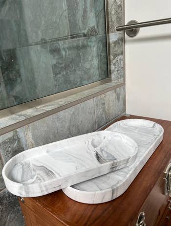 Two oval marble trays are stacked on a wooden surface near a glass shower door