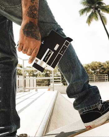 Person with tattooed hand holds vintage camera at skatepark, wearing jeans and sneakers, with palm tree in background