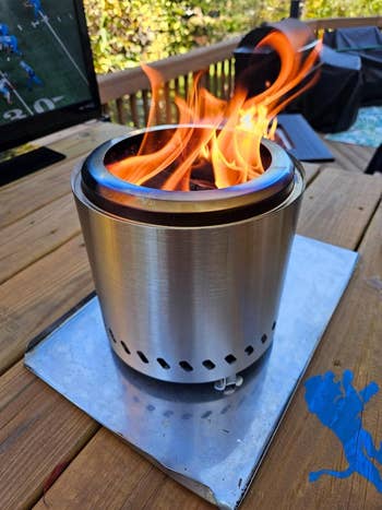Portable metallic  occurrence  pit with flames connected  a woody  deck, adjacent  to an outdoor TV showing a sports game