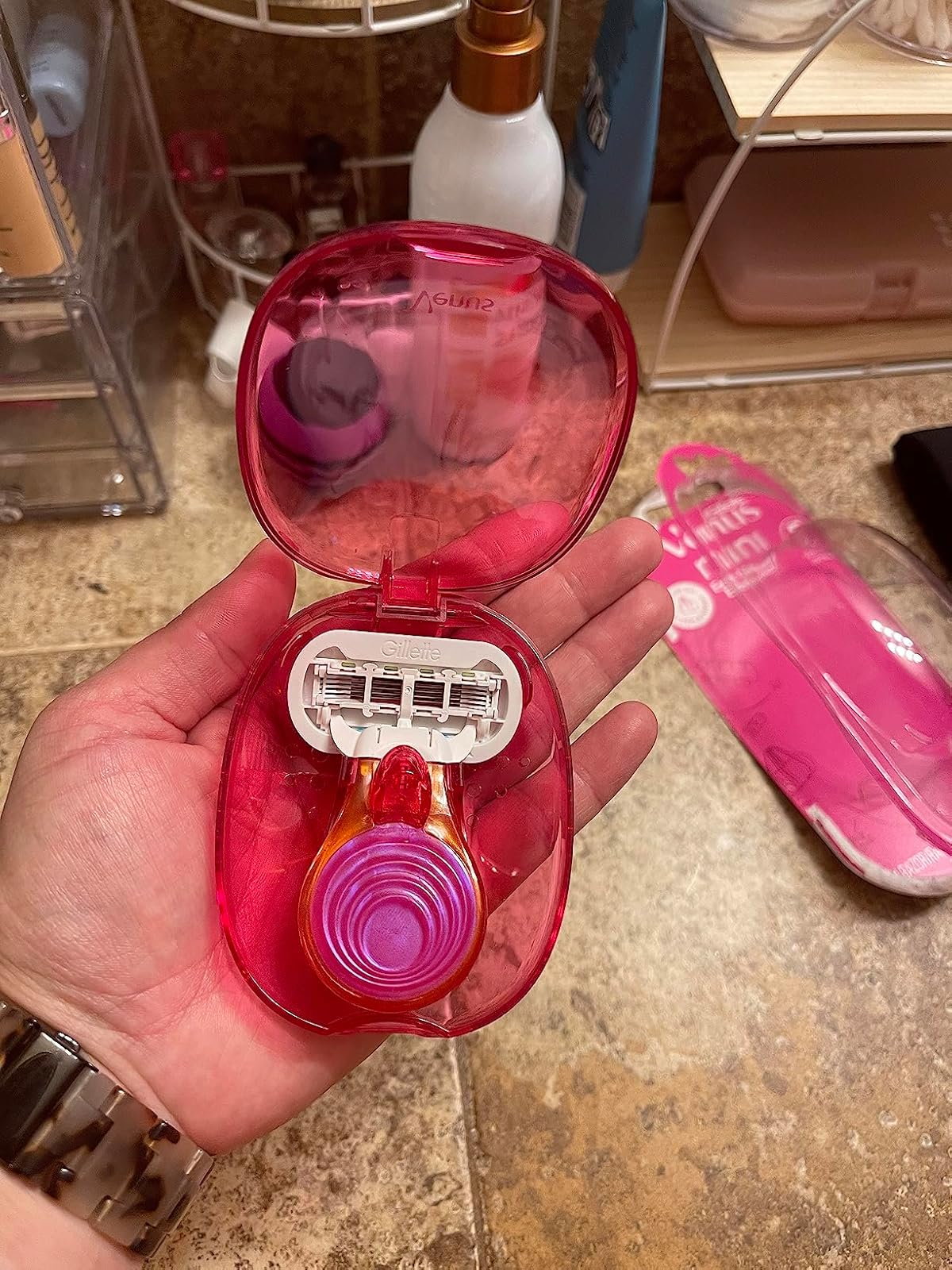 Hand holding a pinkish  razor kit with 3  replacement blades and a matching pinkish  razor case. Background shows assorted  bath  items
