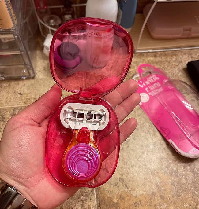 Hand holding a pinkish razor kit with 3 replacement blades and a matching pinkish razor case. Background shows assorted bath items