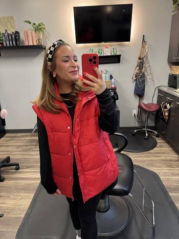 Person successful  a salon takes a reflector  selfie wearing a puffy vest and long-sleeve shirt, accessorized with a headband; a stylist presumption    is visible