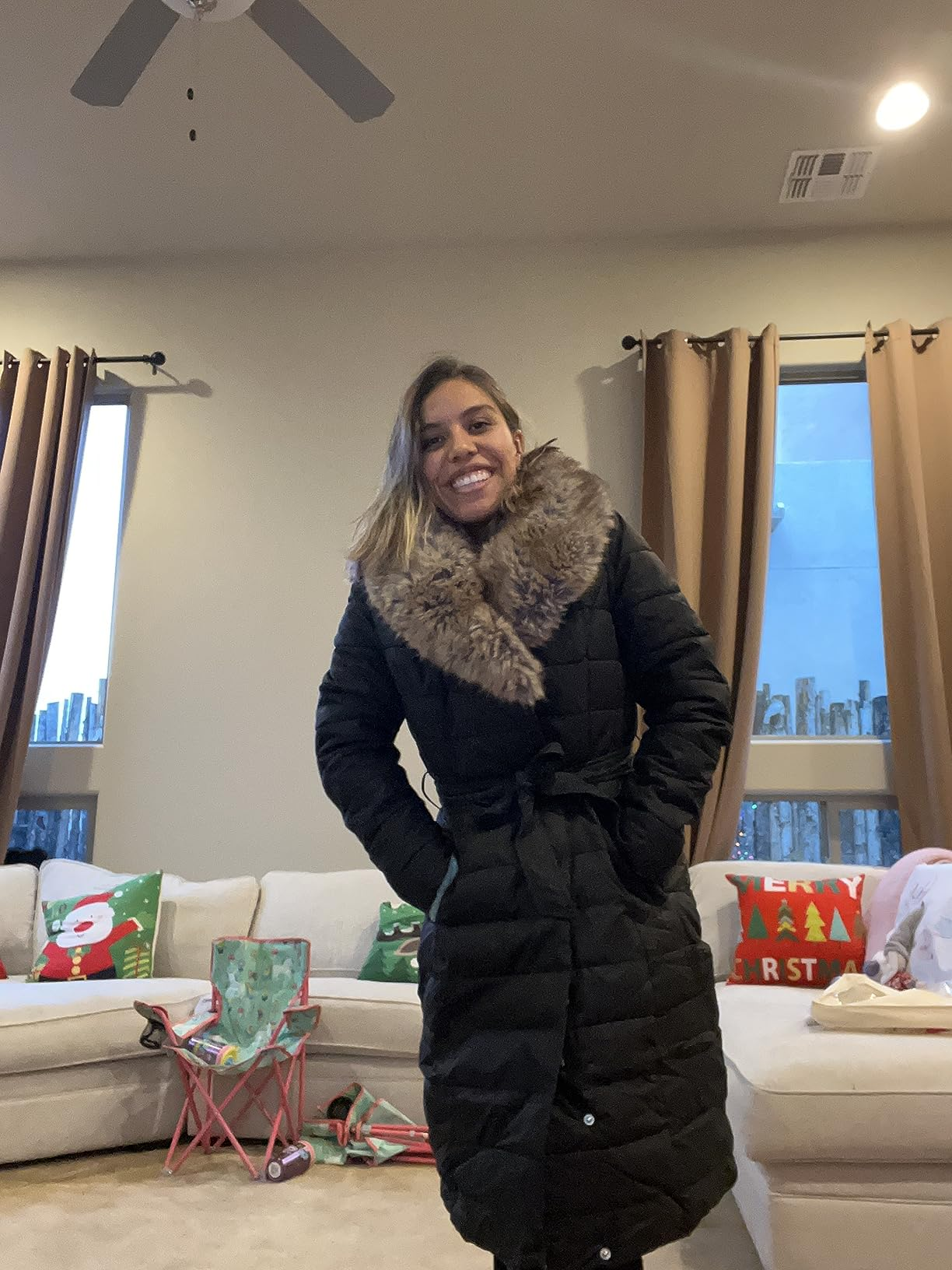 A idiosyncratic   smiling successful  a surviving  room, wearing a agelong  quilted overgarment  with a faux fur collar. Holiday-themed pillows and decor are successful  the background
