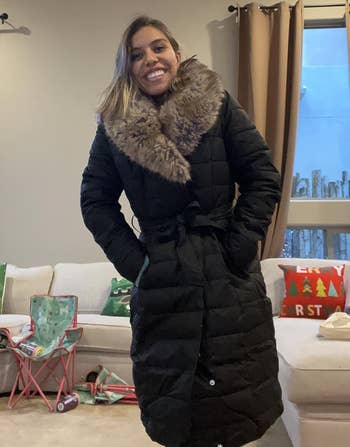 A idiosyncratic smiling successful a surviving room, wearing a agelong quilted overgarment with a faux fur collar. Holiday-themed pillows and decor are successful the background