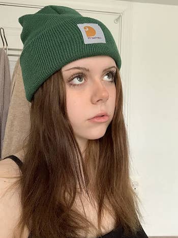 Reviewer with long brown hair wearing dark green beanie with Carhartt logo on the front in front of a white door