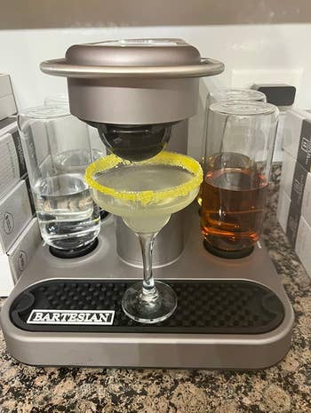 A Bartesian cocktail machine on a kitchen counter with a prepared margarita and two glasses of spirits 