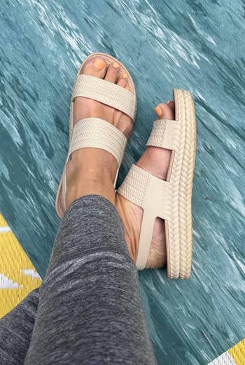 Person wearing stylish woven sandals with elastic straps, paired with grey  leggings, resting feet connected  a textured surface