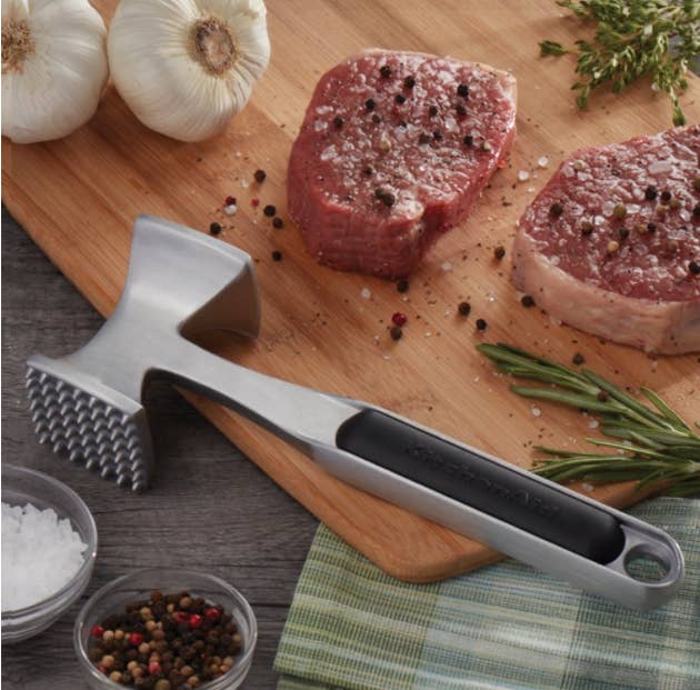 A metallic meat tenderizer on a wooden cutting board next to two slabs of meat and a white onion