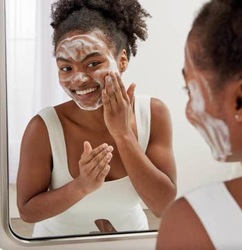 Model smiling and applying foaming facial cleanser while looking in a bathroom mirror