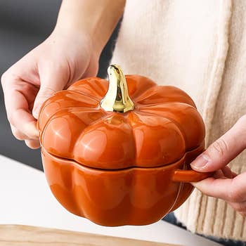 Person holding a pumpkin-shaped ceramic vessel  with a lid and handle, designed for serving food