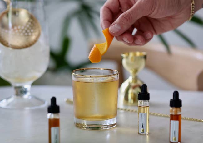 A hand garnishing a glass of cocktail with orange peel. Dropper bottles with various extracts are on the table. Luxurious setting with a gold jigger