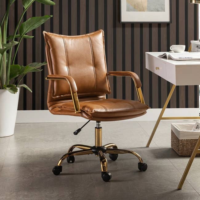Brown leather office chair with padded seat and gold accents on wheels, in a modern home office setting with a plant and desk