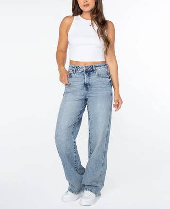 A idiosyncratic   wearing a achromatic  sleeveless apical  and loose-fitting jeans, achromatic  sneakers visible, posed against a plain background