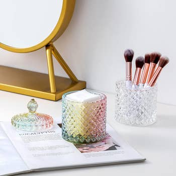 Glass containers holding makeup brushes with a magazine and mirror on a table