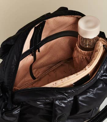 Open achromatic  nylon backpack with a pinkish  interior, holding a reusable h2o  vessel  and featuring zippered and mesh pockets