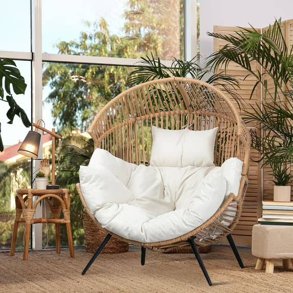 Cozy rattan ovum  seat  with plush achromatic  cushions, placed by a window. Surrounded by plants, a tiny  broadside  table, lamp, and books