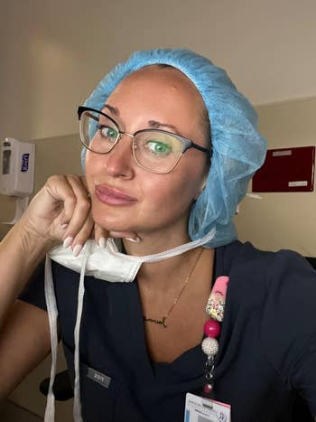 A person in scrubs and a hair net is touching a face mask under their chin, indoors, likely in a medical setting