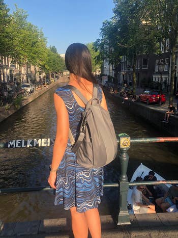 Woman with backpack connected  canal bridge, wearing a wavy patterned dress, facing away. Urban mounting  with radical   successful  a vessel  below