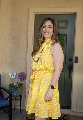 Woman in a patterned yellow sleeveless dress and necklace, smiling on a house porch