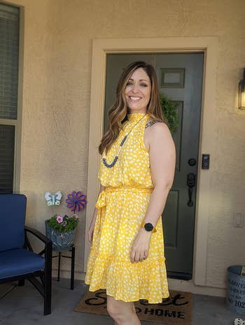 Woman in a patterned yellow sleeveless dress and necklace, smiling on a house porch