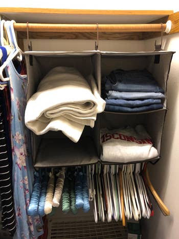 Closet enactment with hanging cloth shelves holding folded jeans, a achromatic blanket, and a printed sweatshirt. Dresses and shirts bent below
