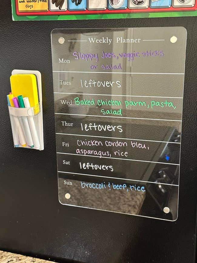 A weekly planner on a refrigerator with meals listed: Monday - Sloppy Joes, veggie sticks or salad; Tuesday, Thursday, and Saturday - leftovers; Wednesday - Baked chicken parm, pasta, salad; Friday - Chicken cordon bleu, asparagus, rice; Sunday - Broccoli and beef, rice. There’s a pen holder with colorful markers next to it