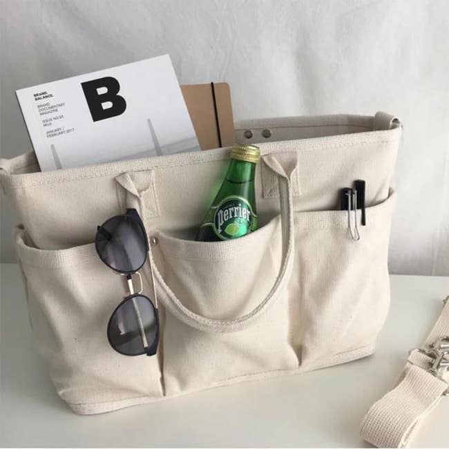 the canvas tote with exterior pockets holding sunglasses, a bottle of water, pens, papers, and a notebook