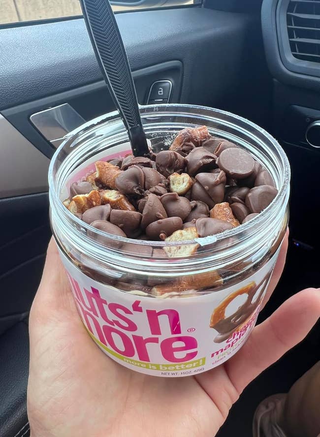 A hand holds a jar of Nuts 'n More, with chocolate-covered pretzel pieces visible on top of the spread