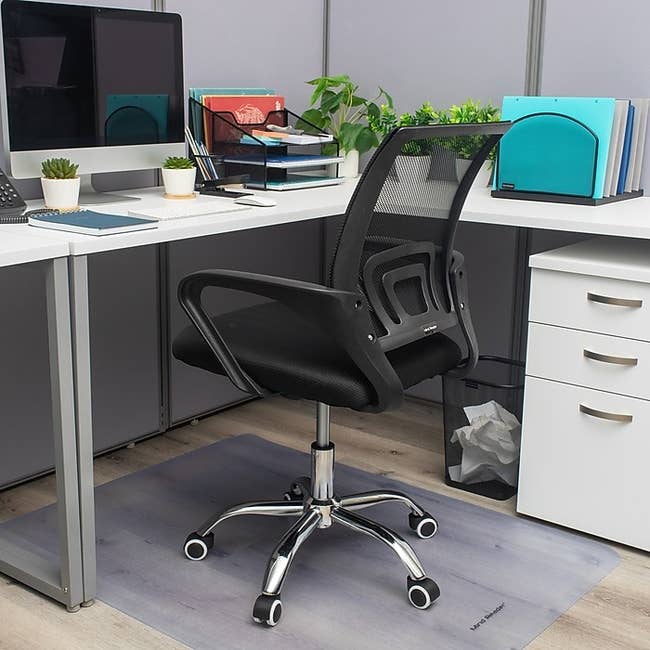 Office chair with mesh back on a mat, positioned at a modern desk setup including a computer, plants, and organized office supplies