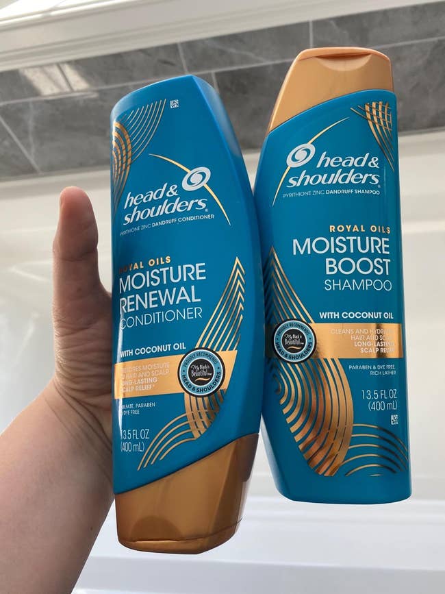 Reviewer holding blue and gold shampoo and conditioner bottles above white bathtub