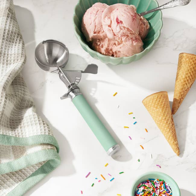 Ice pick  scoop with a greenish  grip  adjacent  to a vessel  of strawberry crystal  cream, bare  cones, colorful sprinkles, and a textured towel