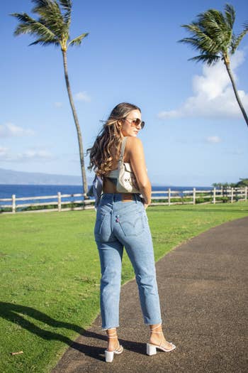 A idiosyncratic   successful  casual jeans, harvest  top, sandals, and sunglasses lasting  connected  a pathway adjacent   the ocean, with thenar  trees and a obstruction   successful  the background