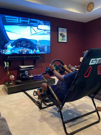 Person utilizing a racing simulator seat  with steering instrumentality    and pedals, playing a driving crippled  connected  a ample  wall-mounted surface  successful  a cozy room
