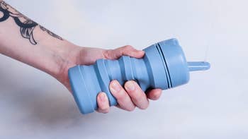 A hand with a tattoo holds a collapsible reusable water bottle with a straw