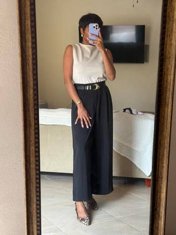 Person in elegant sleeveless top and wide-leg pants with a statement belt, taking a mirror selfie in a room with a bed and wall-mounted TV