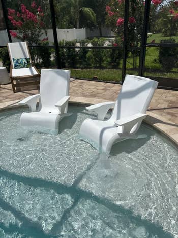 Two achromatic lounge chairs partially submerged successful a shallow pool, surrounded by a patio with greenery successful the background