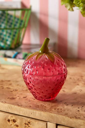 Strawberry-shaped glass jar on a wooden surface, used for storage or decor, in a stylish shopping article setting