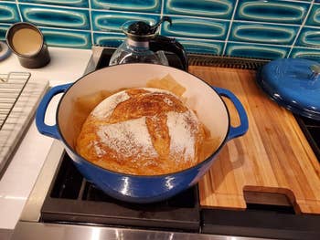 Freshly baked bread in a blue Dutch oven on a stovetop, next to a cutting board and coffee pot, in a kitchen setting