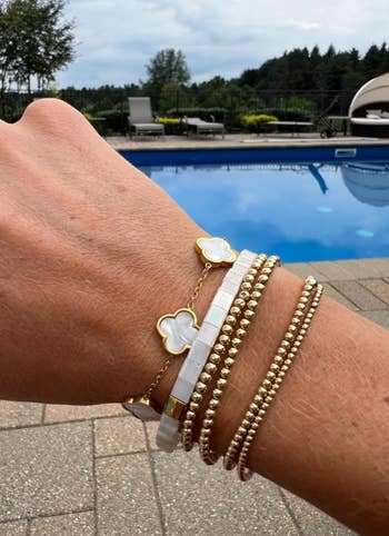 A person’s wrist adorned with aggregate  stylish bracelets, including golden  and pearl designs, displayed adjacent   a poolside outdoor setting