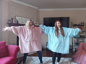 Two people happily pose in oversized, cozy wearable blankets with large front pockets in a living room setting