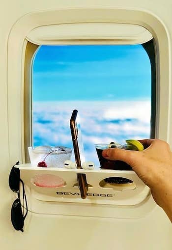 A person's hand holding lifting a drink out of a flight tray that also has their phone, another drink, and their sunglasses on it near a window on a plane