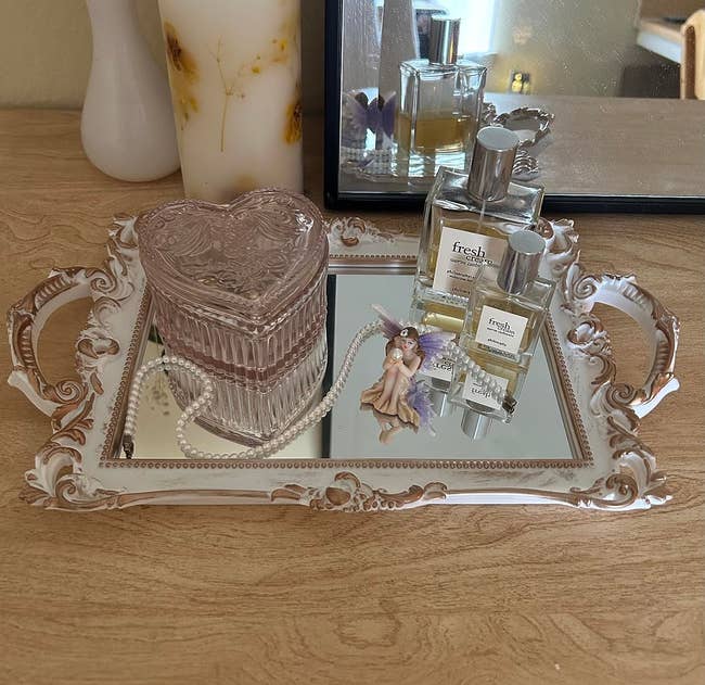 A dresser with a mirror tray holding perfumes, a heart-shaped box, and a fairy figurine