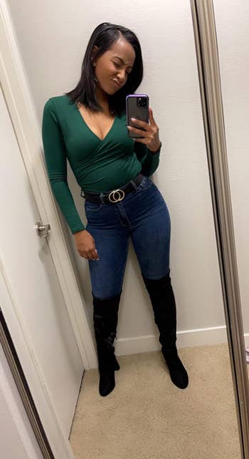 A reviewer taking a mirror selfie, wearing a fitted green wrap top, blue jeans, a black belt with a gold buckle, and black knee-high boots